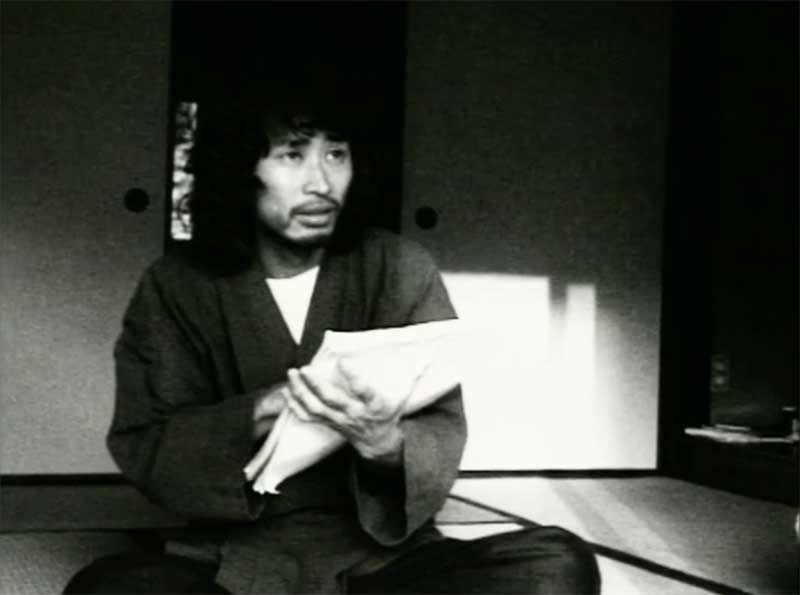 image en noir et blanc, présentant un homme japonais en tenue traditionnelle, en position assise, dessine sur sur du papier blanc